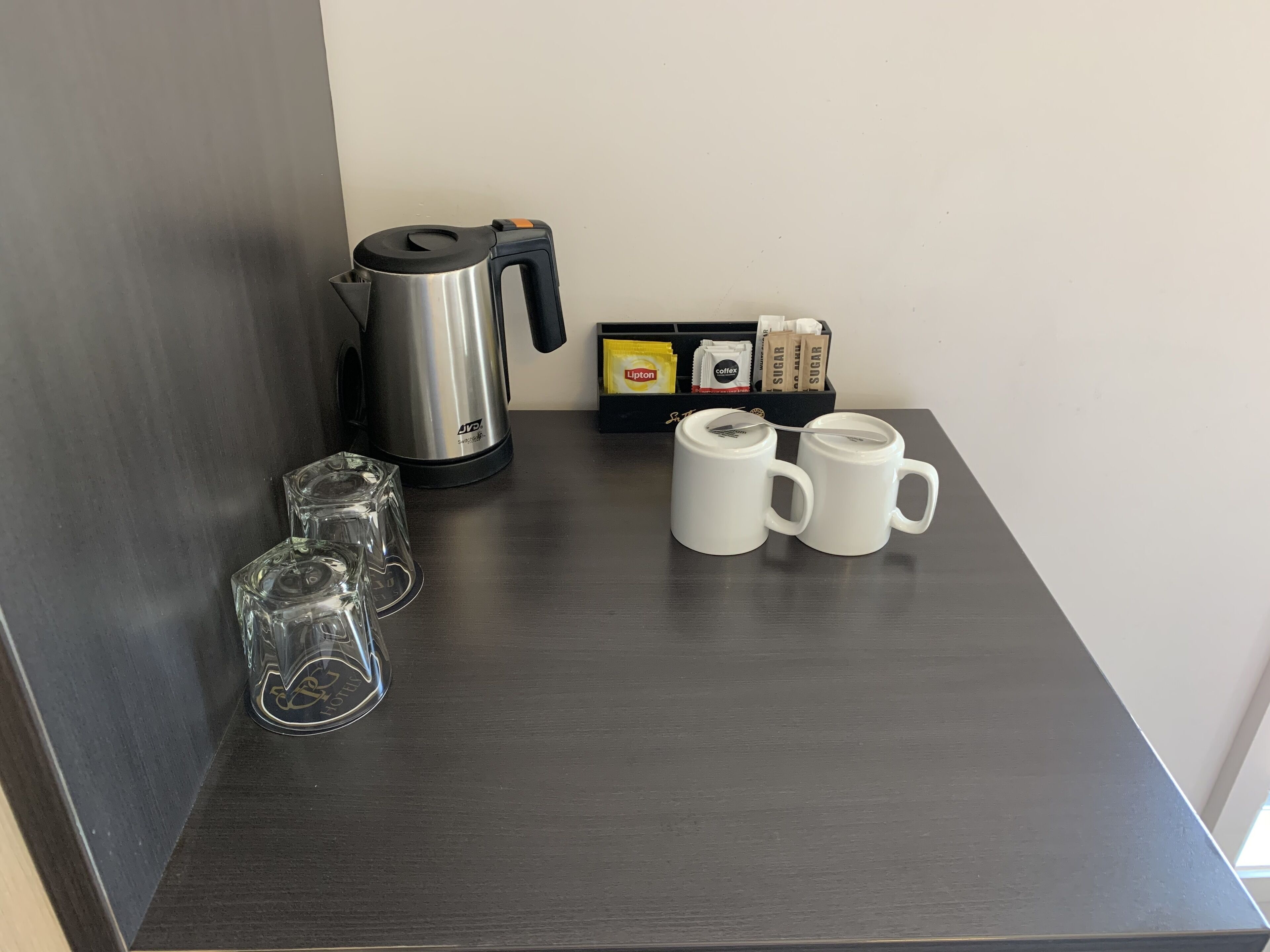 A kitchen counter with a coffee maker, two mugs, a glass, and sugar packets.