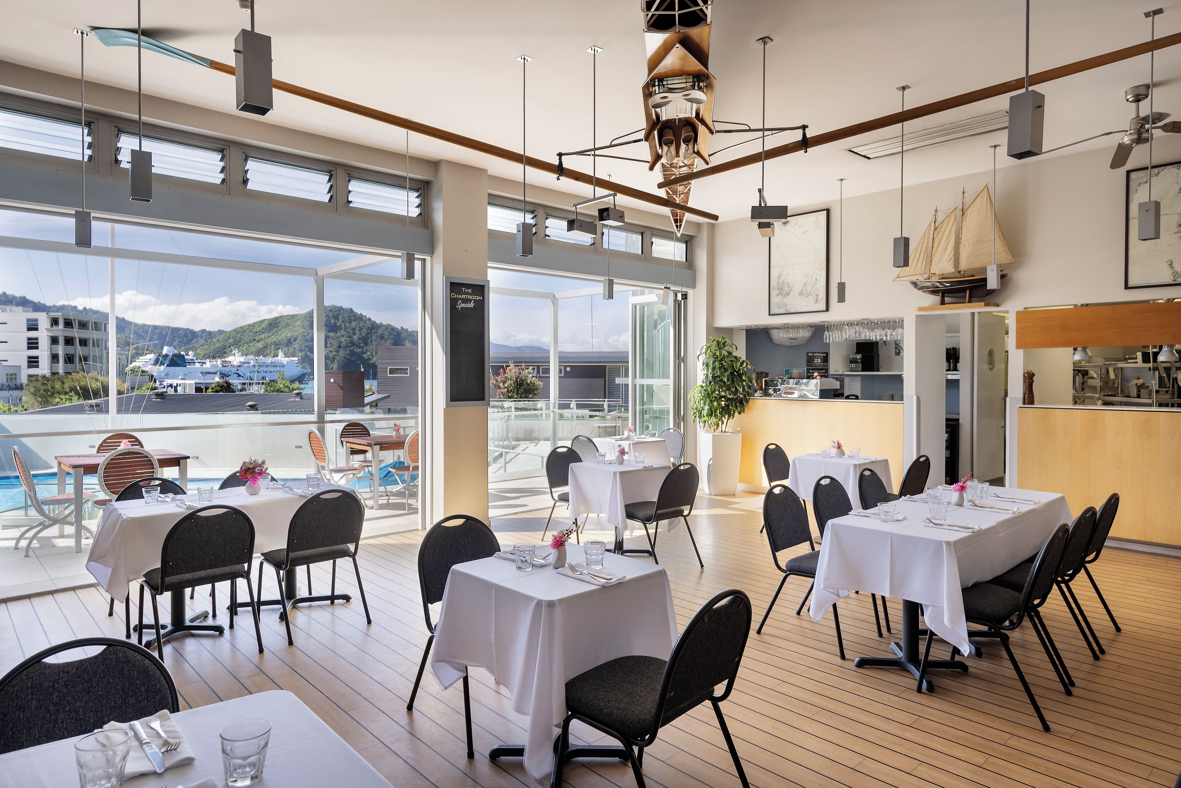 En restaurang med många bord och stolar, stora fönster med utsikt över havet och båtar, och en modern inredning.