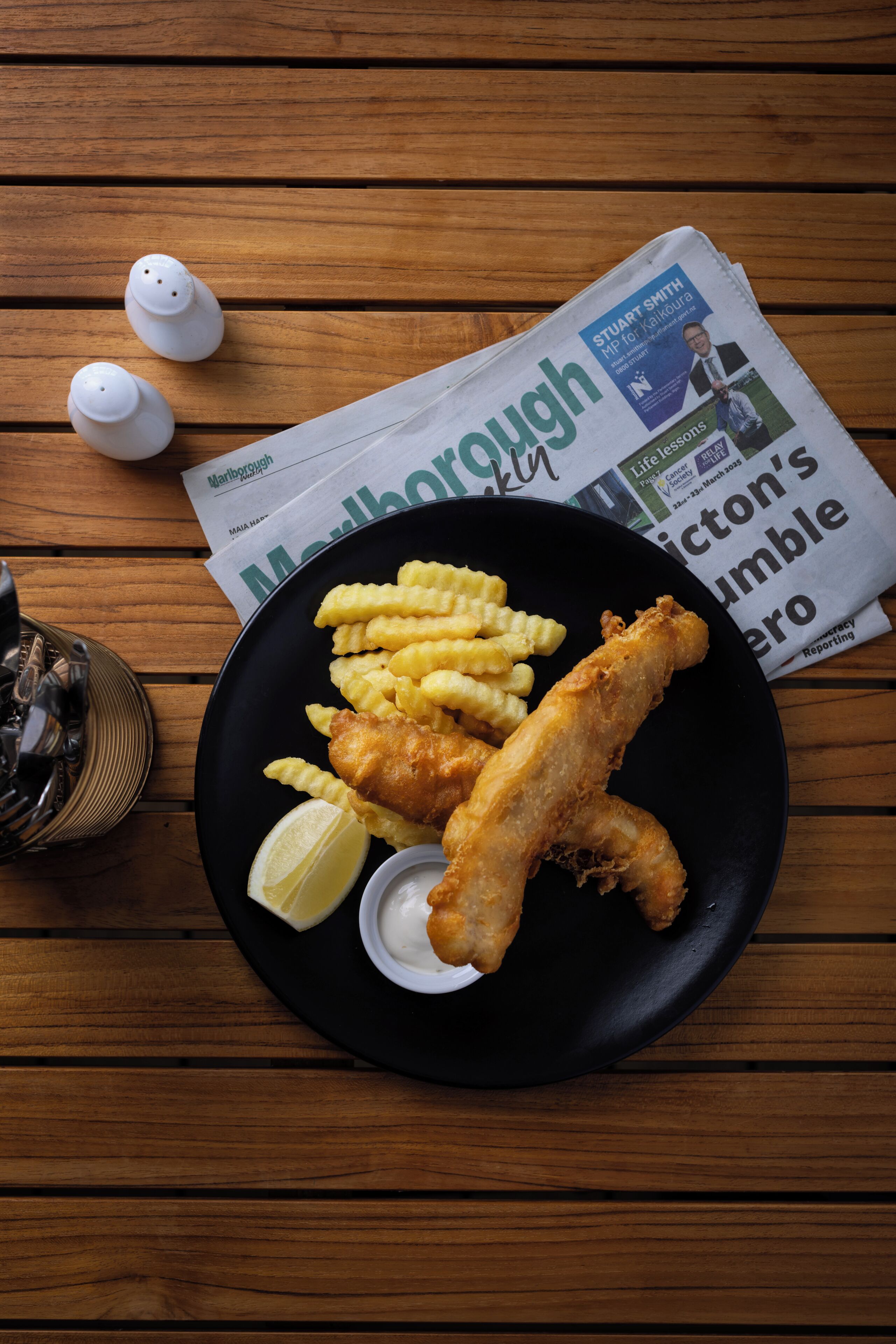 A plate of fish and chips with lemon wedges and sauce, a newspaper, and salt and pepper shakers on a wooden table.