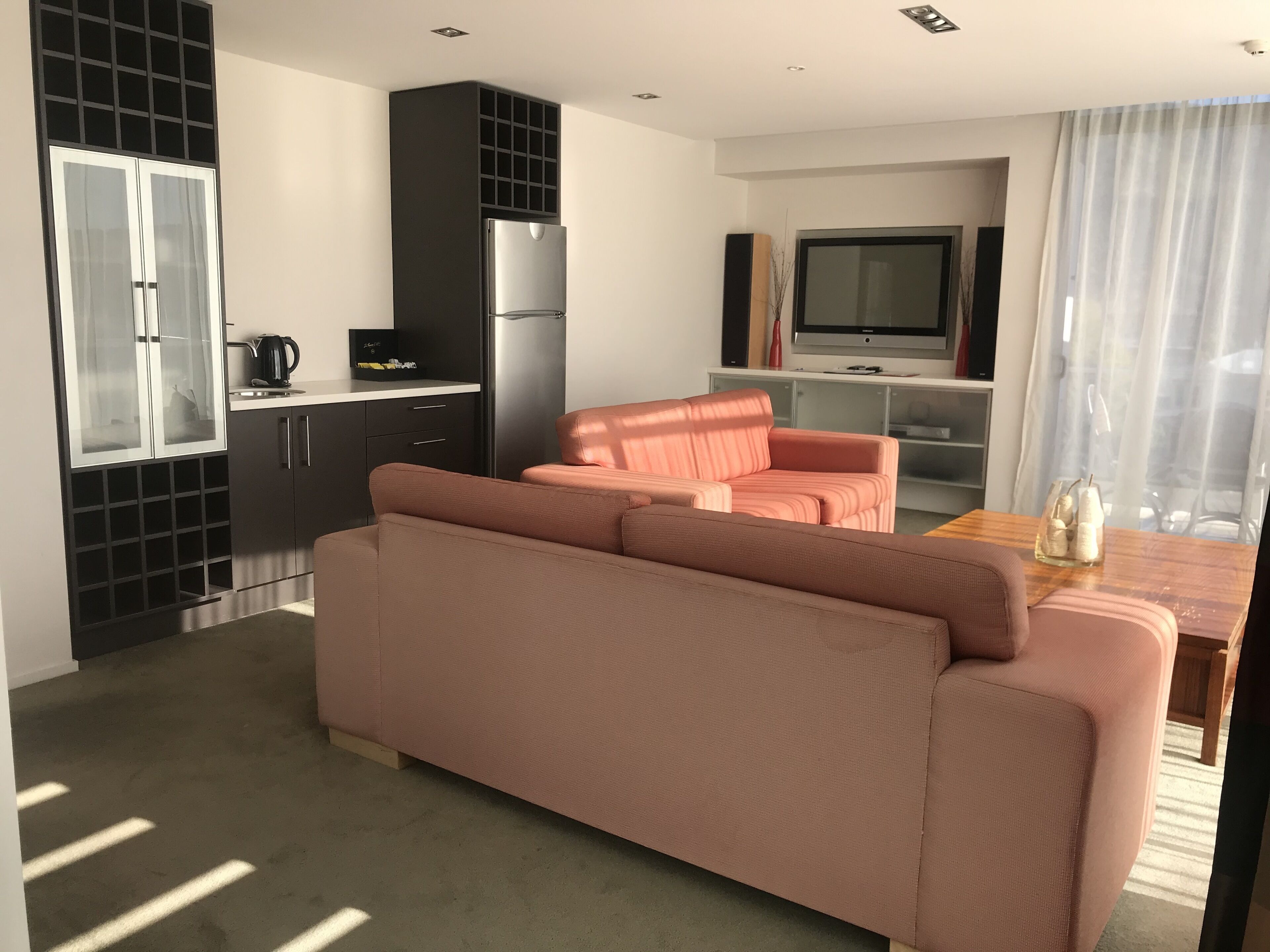 A modern living room with a pink sofa, a kitchen area with a black refrigerator, and a dining table with a vase of flowers.