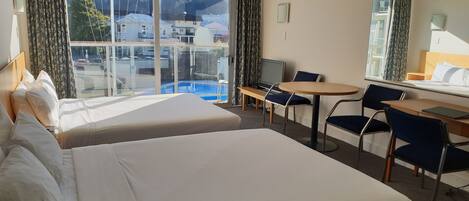 A hotel room with two beds, a desk, a chair, a TV, and a view of a swimming pool and buildings.