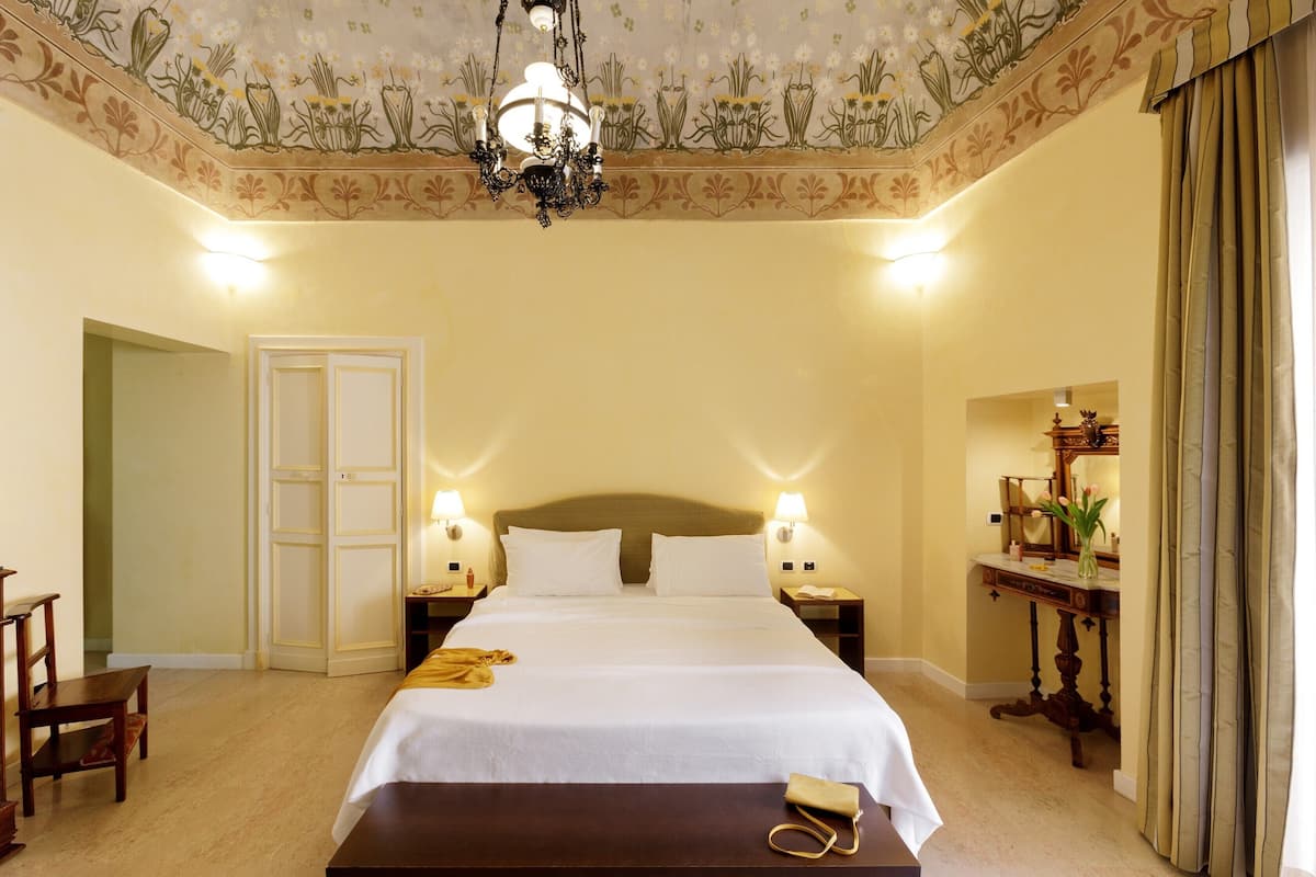 A hotel room with a large bed, two bedside tables with lamps, a wooden nightstand, a desk with a chair, and a mirror.