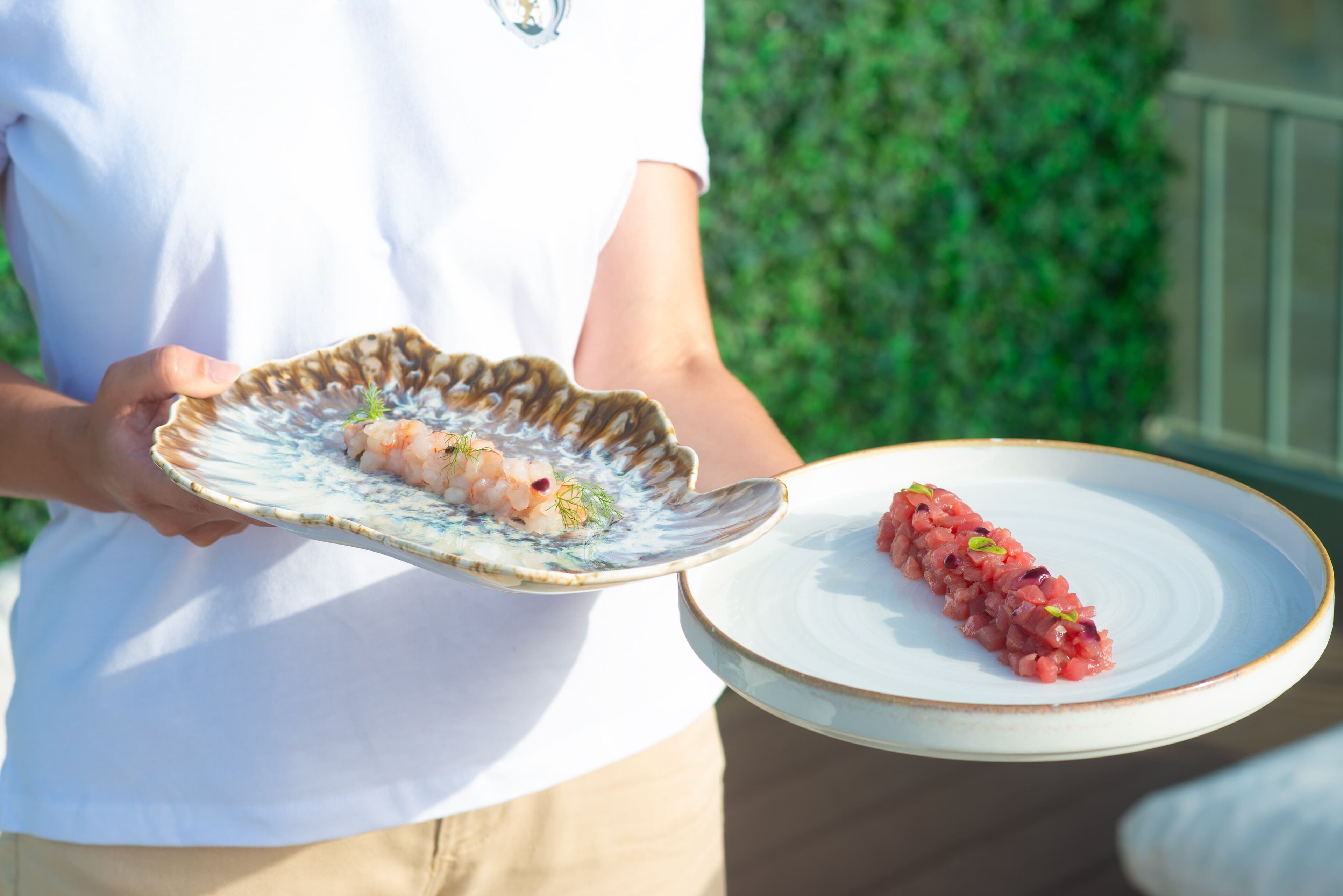 A person holding two plates of food, one with shrimp and the other with a red dish, outdoors.