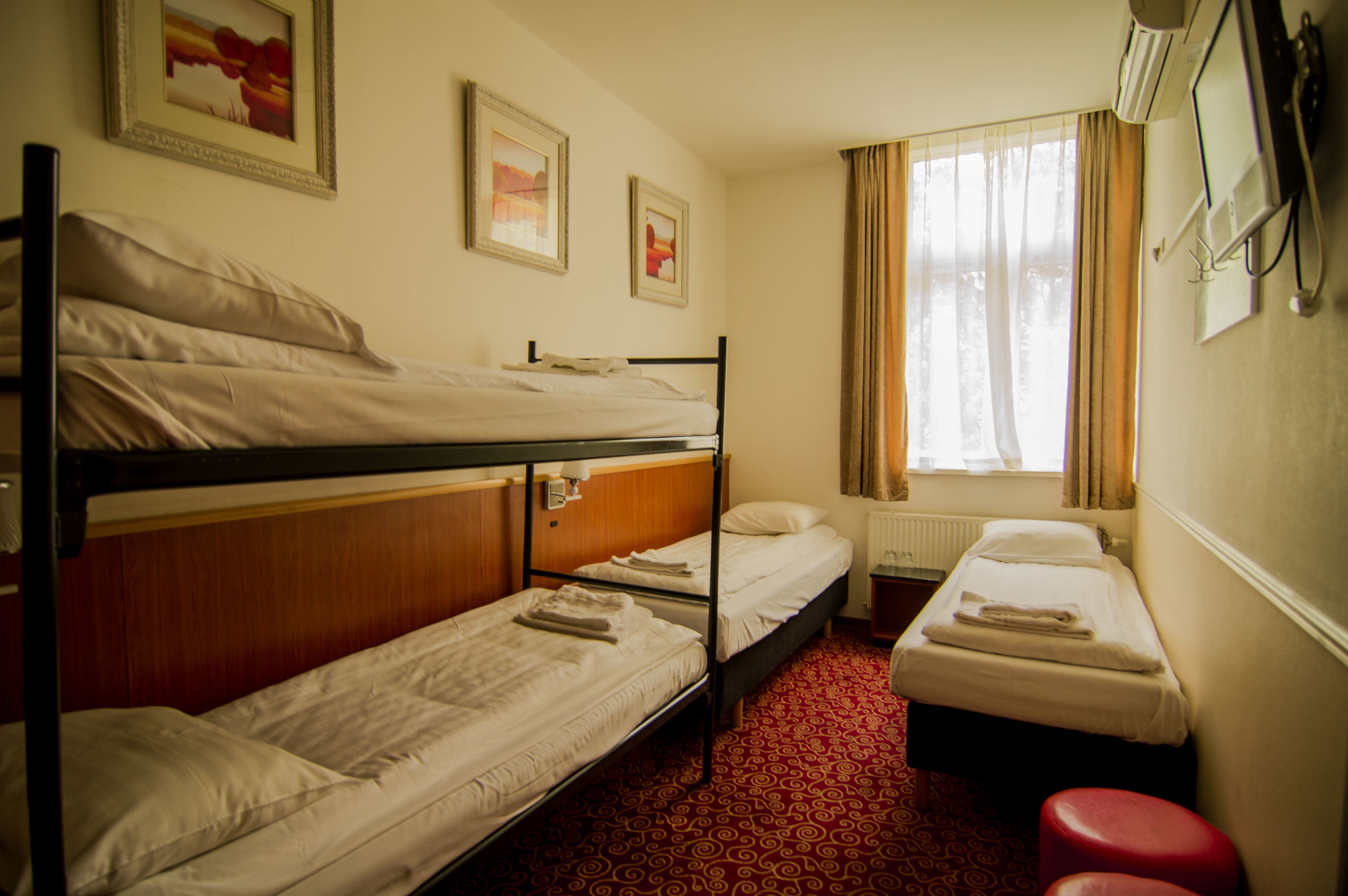 A hotel room with bunk beds, a small single bed, a red patterned carpet, a window with curtains, and a mounted TV.