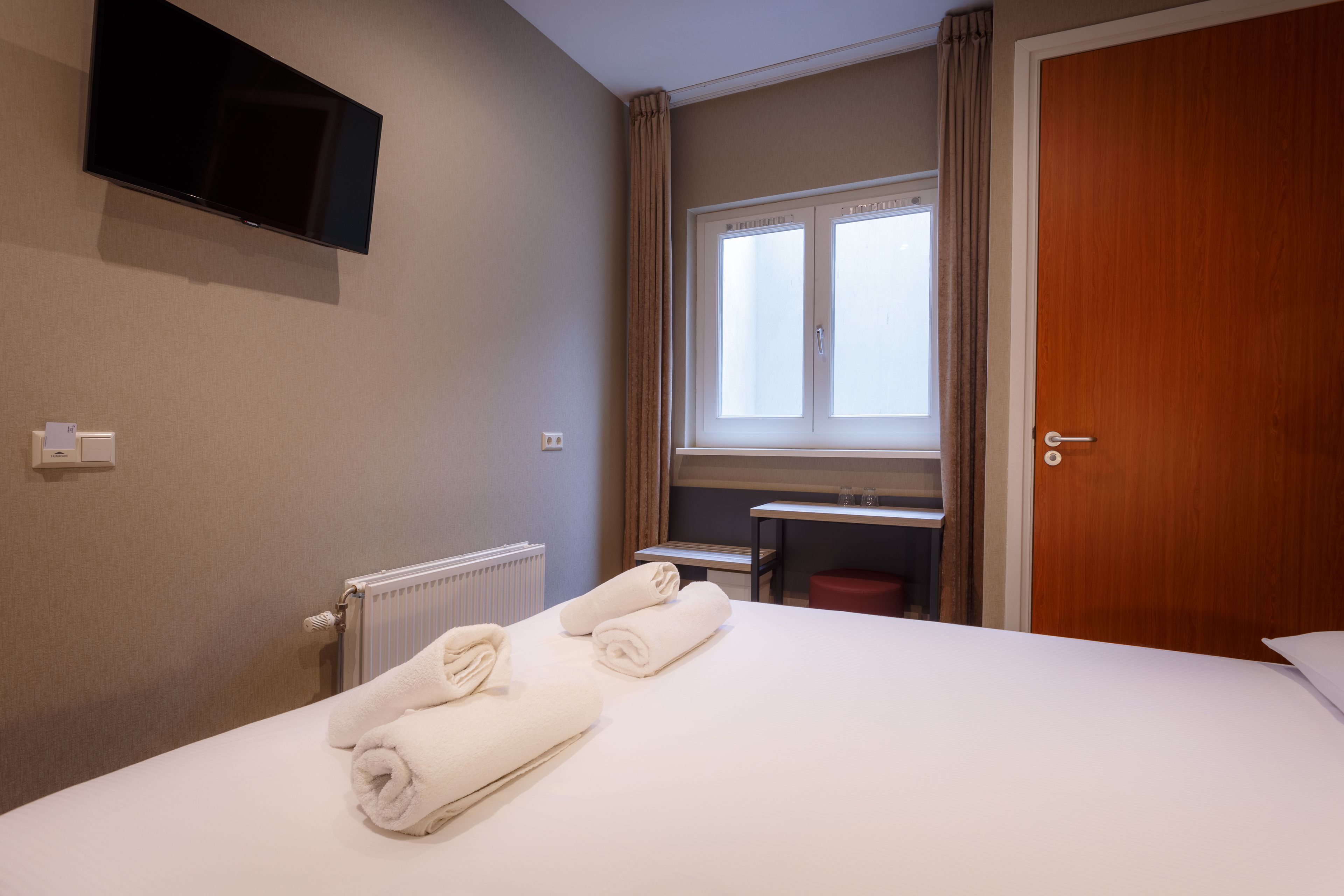 A hotel room with a bed, a TV mounted on the wall, a window with curtains, and a door.