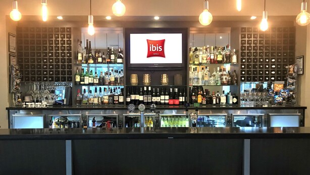 Zona de bar con una pantalla grande mostrando el logotipo de ibis, estantes llenos de diferentes botellas y lámparas colgantes.
