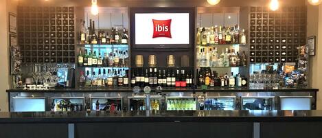Zona de bar con una pantalla grande mostrando el logotipo de ibis, estantes llenos de diferentes botellas y lámparas colgantes.