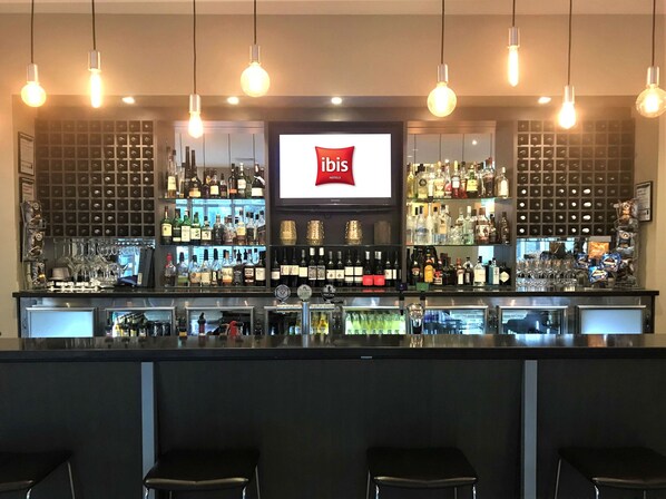 A bar area with a large screen displaying the ibis logo, shelves stocked with various bottles, and hanging light bulbs. - ibis Melbourne Glen Waverley (Glen Waverley)