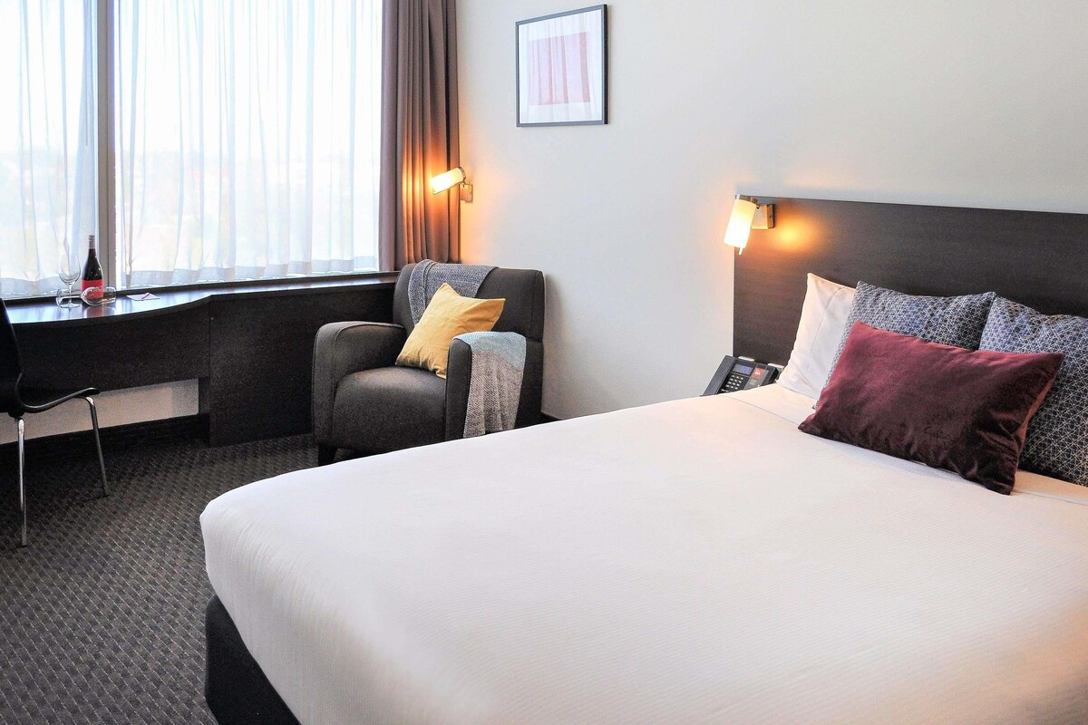 Habitación de hotel con cama, escritorio, silla, teléfono y ventana con cortinas.