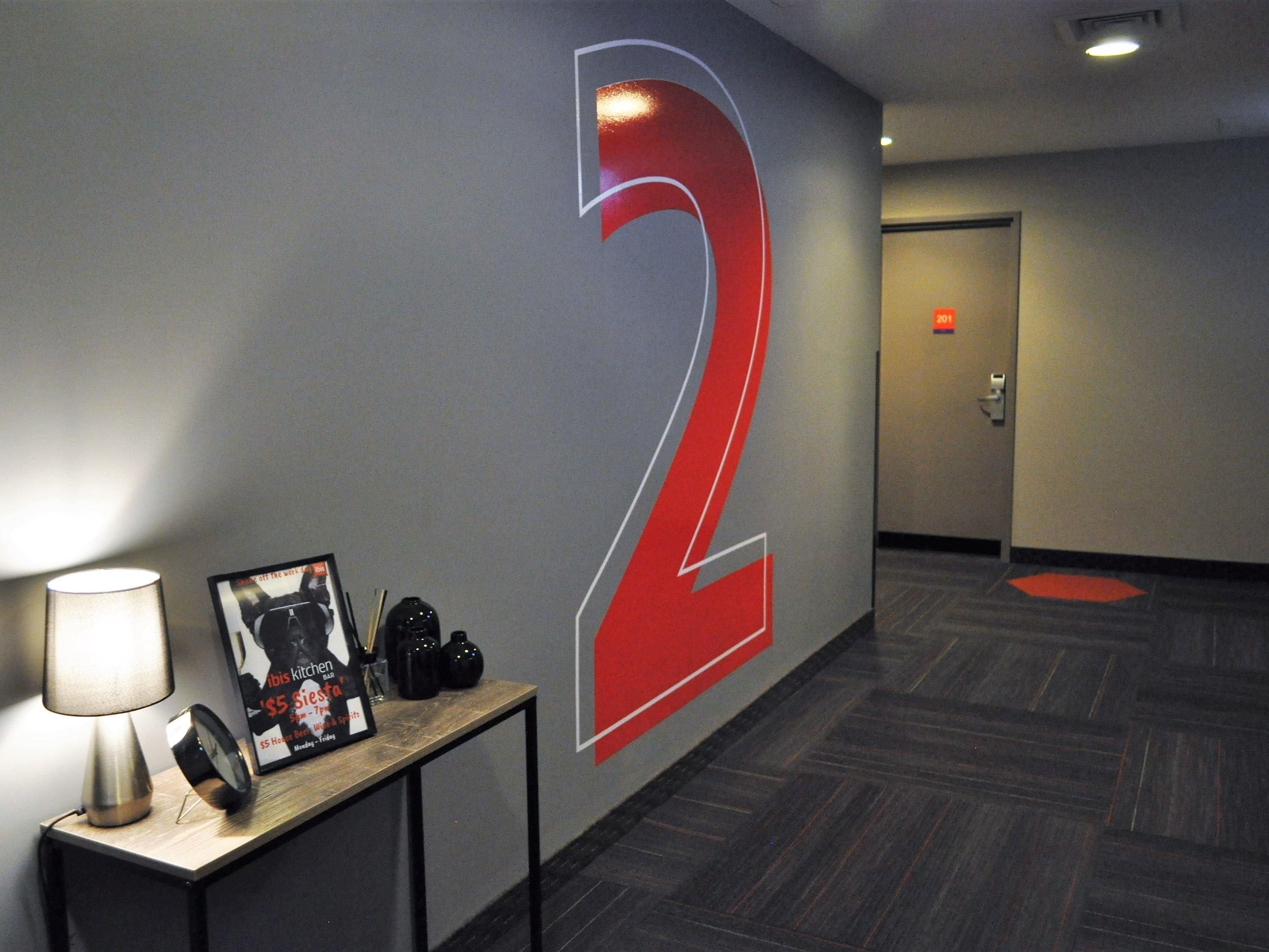 A hallway with a large illuminated number 2 on the wall, a table with a lamp, and a framed picture.