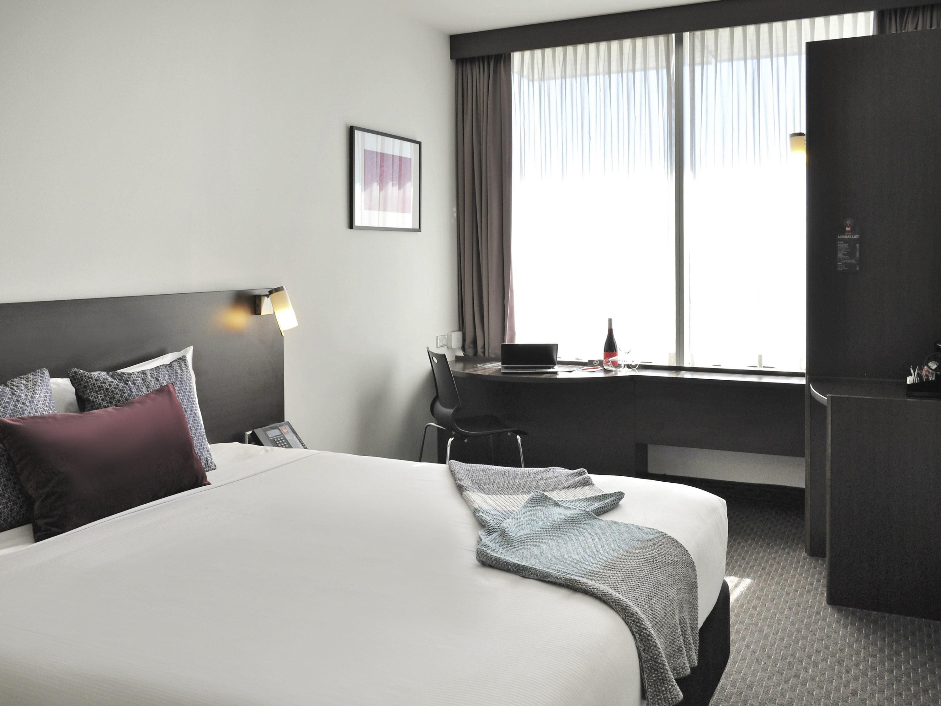 Una habitación de hotel con una cama grande, un escritorio con una computadora portátil, una silla y un refrigerador.