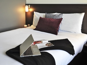 Una cama bien hecha con un ordenador portátil, un mando a distancia y una revista encima.