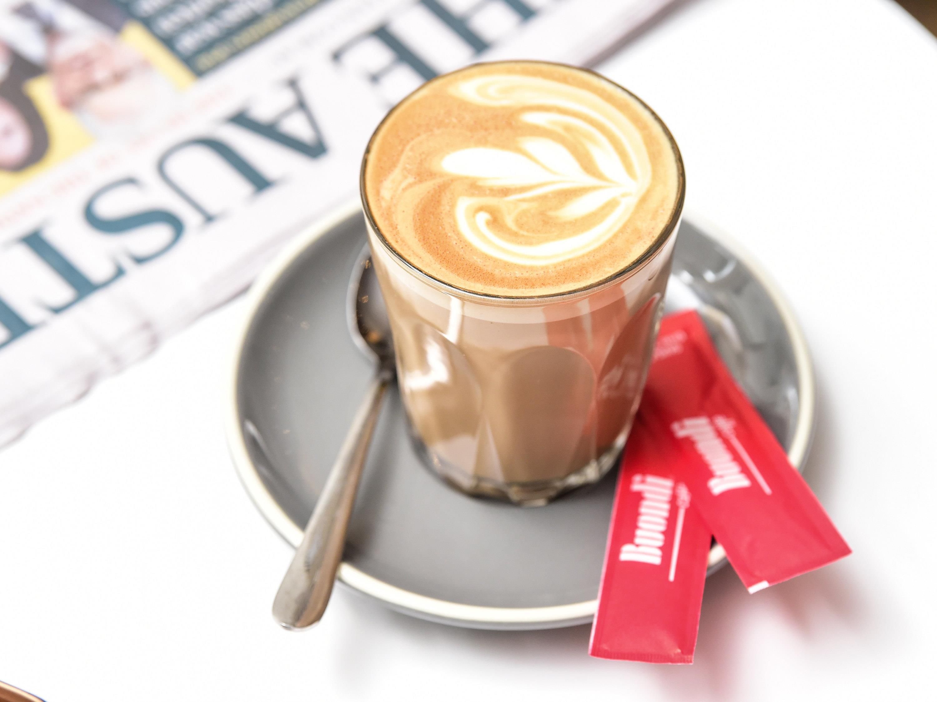 Een kop koffie met latte-art op een schotel, een lepel en een rood zakje ernaast.