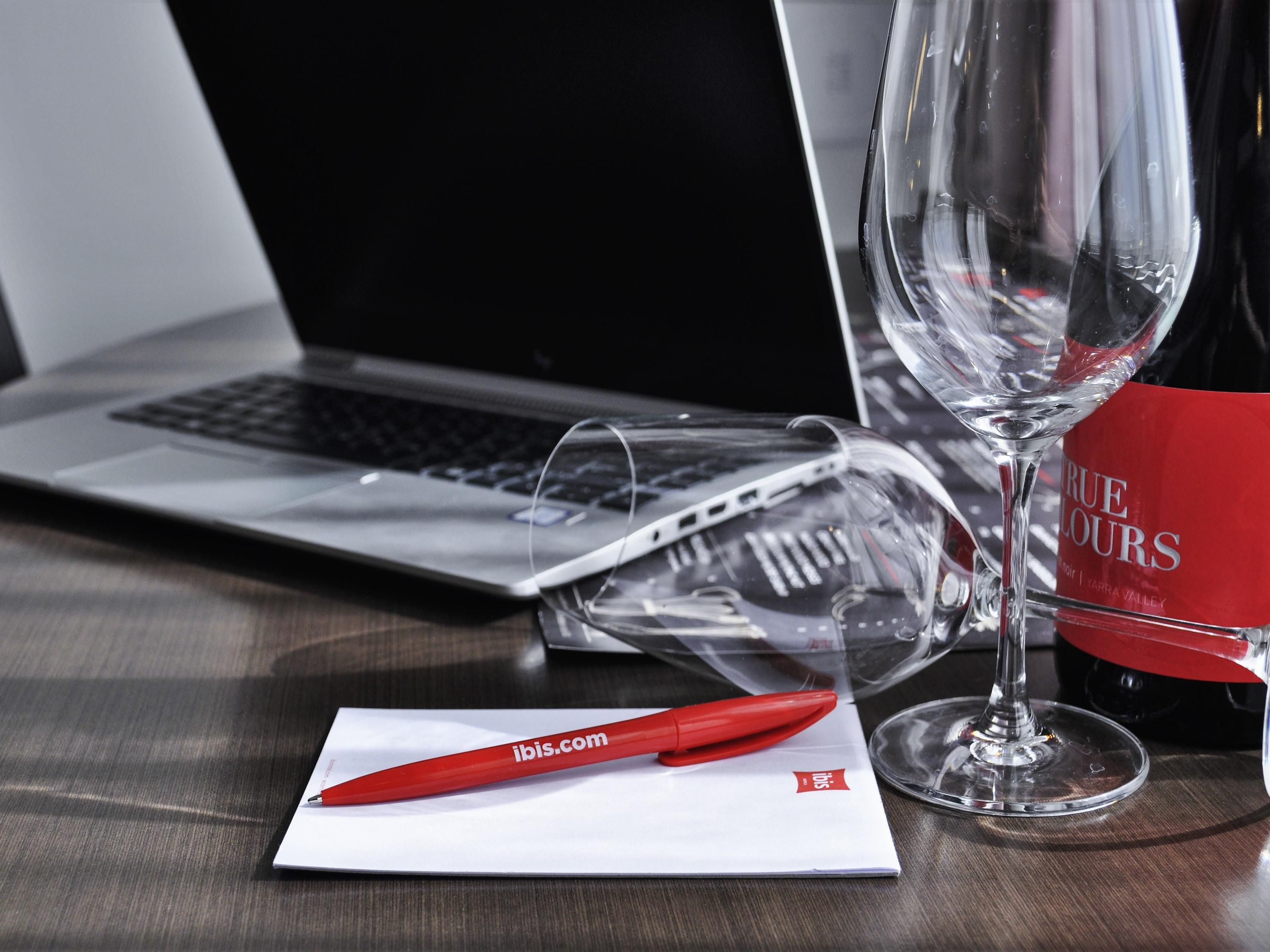 A laptop, a wine glass, a red pen, and a brochure on a wooden table.