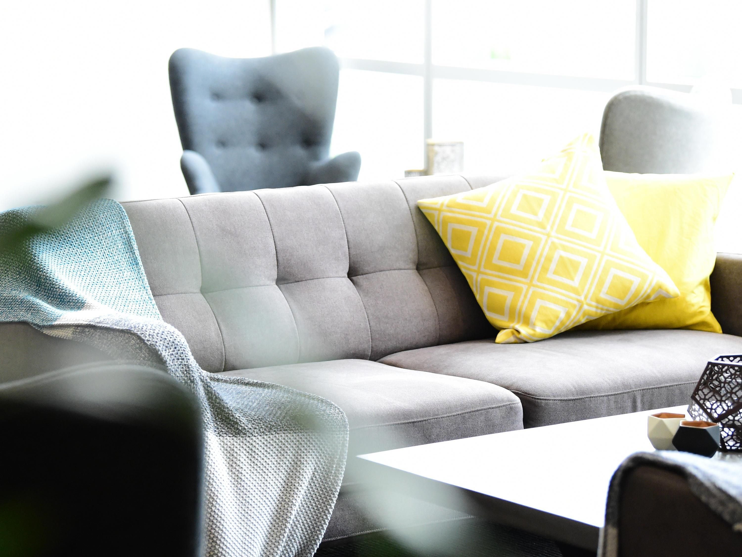 Un sofá gris con un cojín amarillo estampado, un sillón gris y una mesa blanca con un jarrón de diseño geométrico.