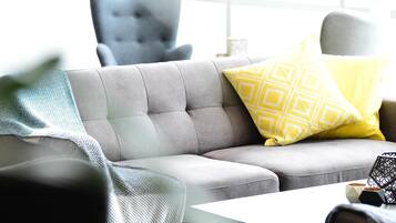 Un sofá gris con un cojín amarillo estampado, un sillón gris y una mesa blanca con un jarrón de diseño geométrico.