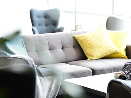 A grey sofa with a yellow patterned pillow, a grey armchair, and a white table with a geometric vase.