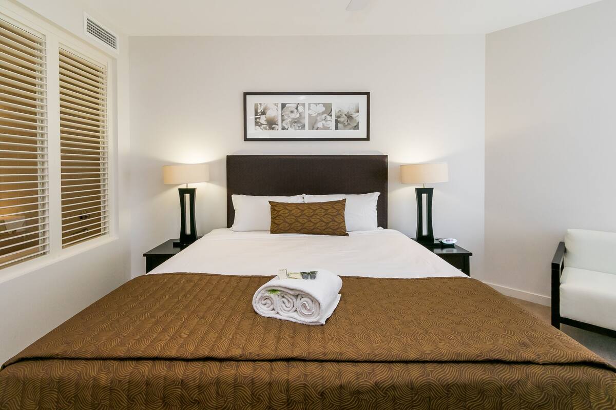 A hotel room with a neatly made bed, a brown patterned bedspread, a framed floral artwork above the bed, two bedside lamps, and a white chair.