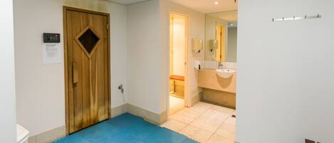 Pasillo de hotel con puerta de madera, felpudo azul, baño con lavamanos y espejo, y un banco.