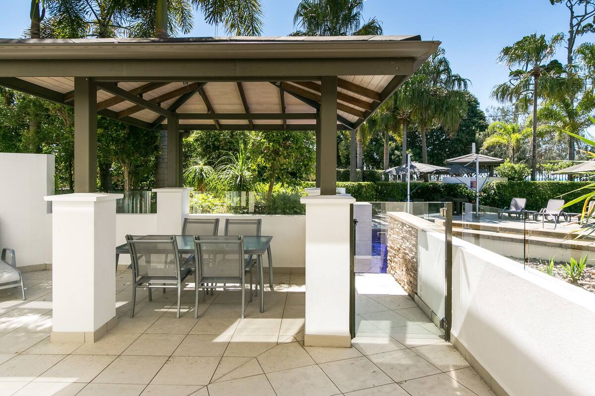 A covered outdoor seating area with a pool view, surrounded by palm trees and greenery.