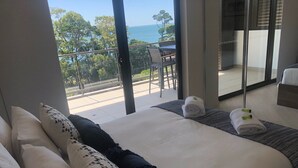 Un dormitorio con una cama grande, dos almohadas, una toalla enrollada y vista al mar desde un balcón.