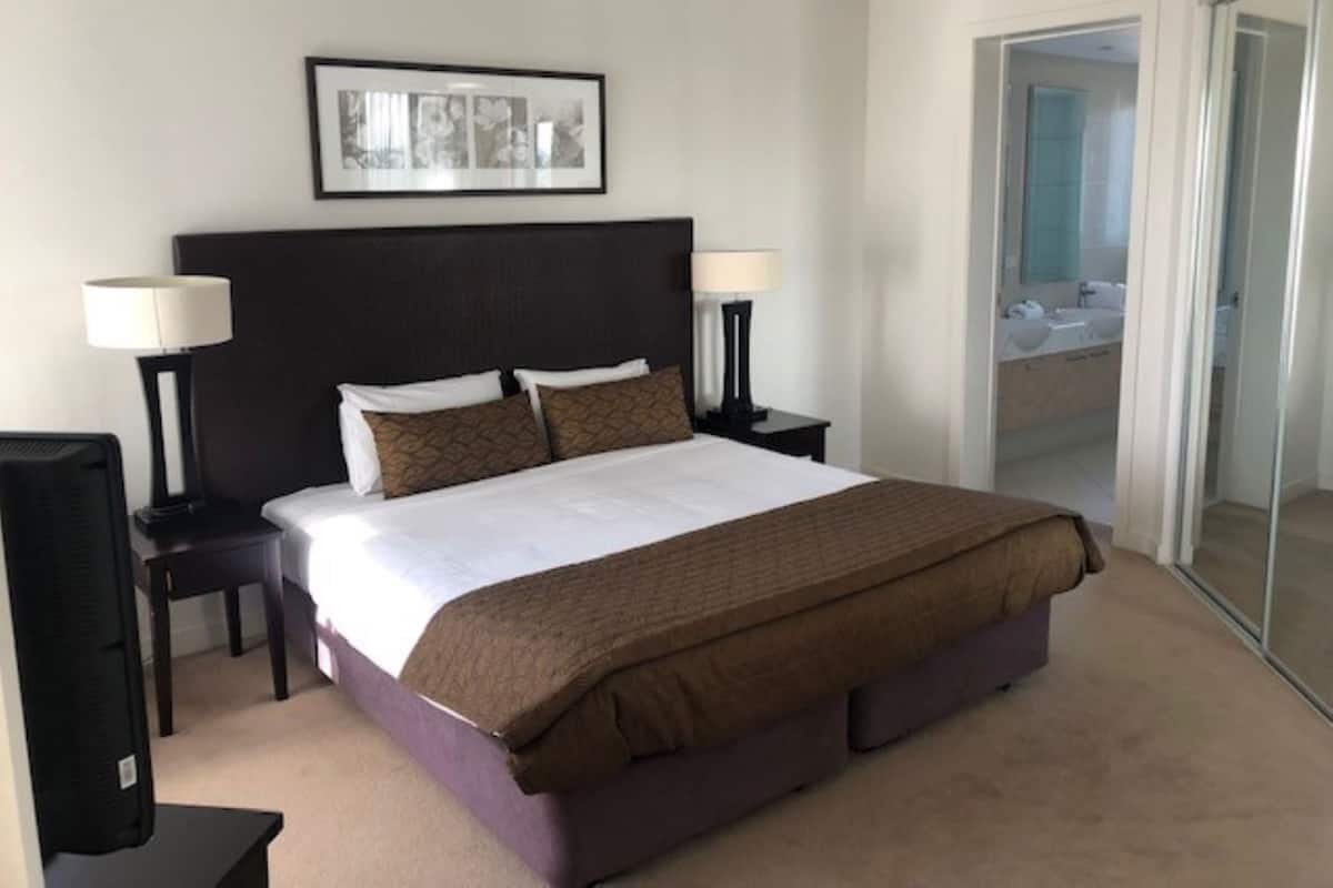 A hotel room with a large bed, two bedside lamps, a ceiling fan, a framed picture on the wall, and a bathroom visible through a glass door.