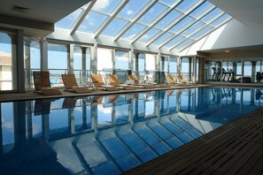 A large indoor swimming pool with clear glass walls, wooden deck, and sun loungers. - WOW Istanbul Hotel (Istanbul)