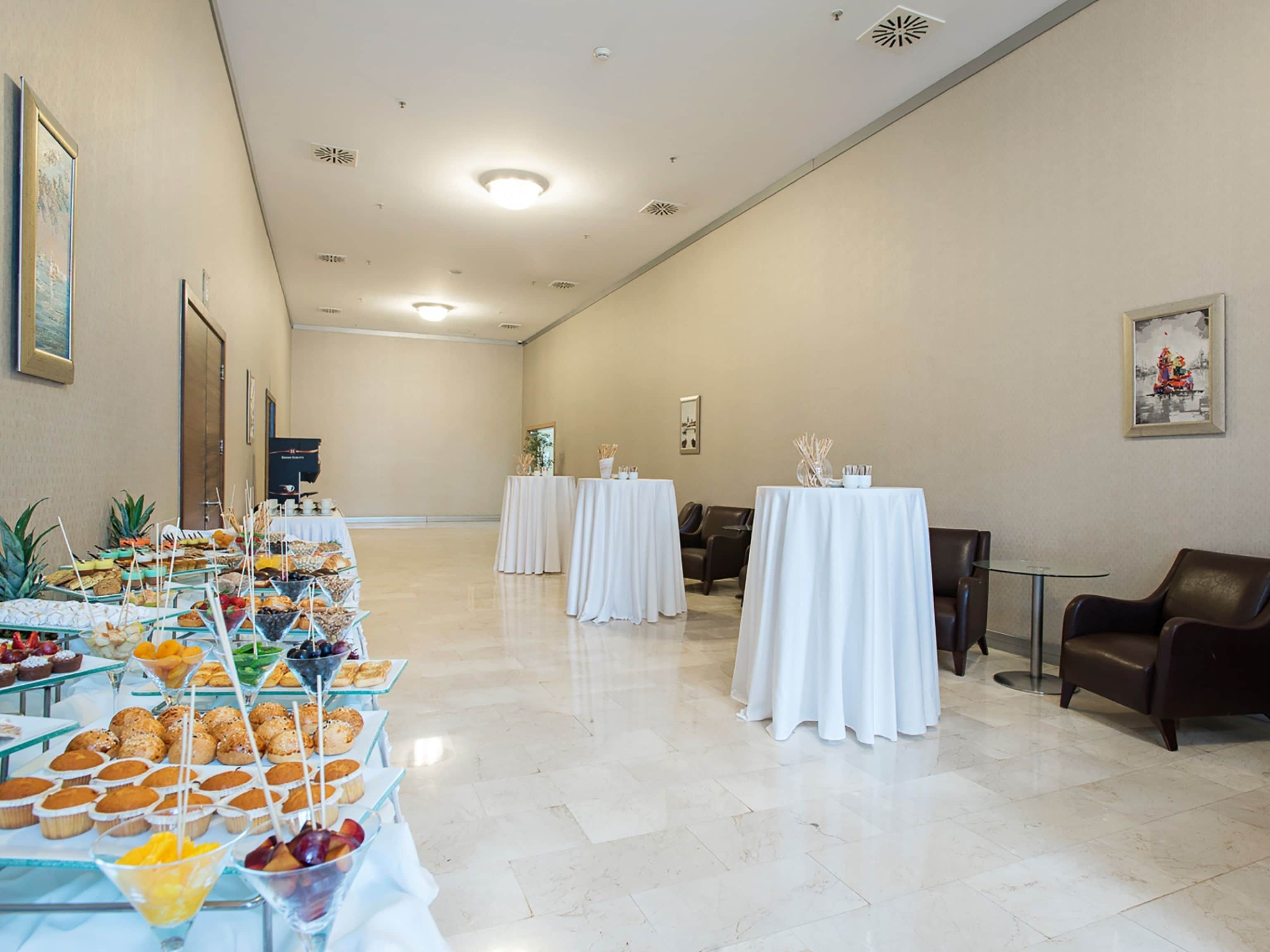 A spacious room with a long buffet table filled with various snacks and drinks, and several smaller tables with white tablecloths.