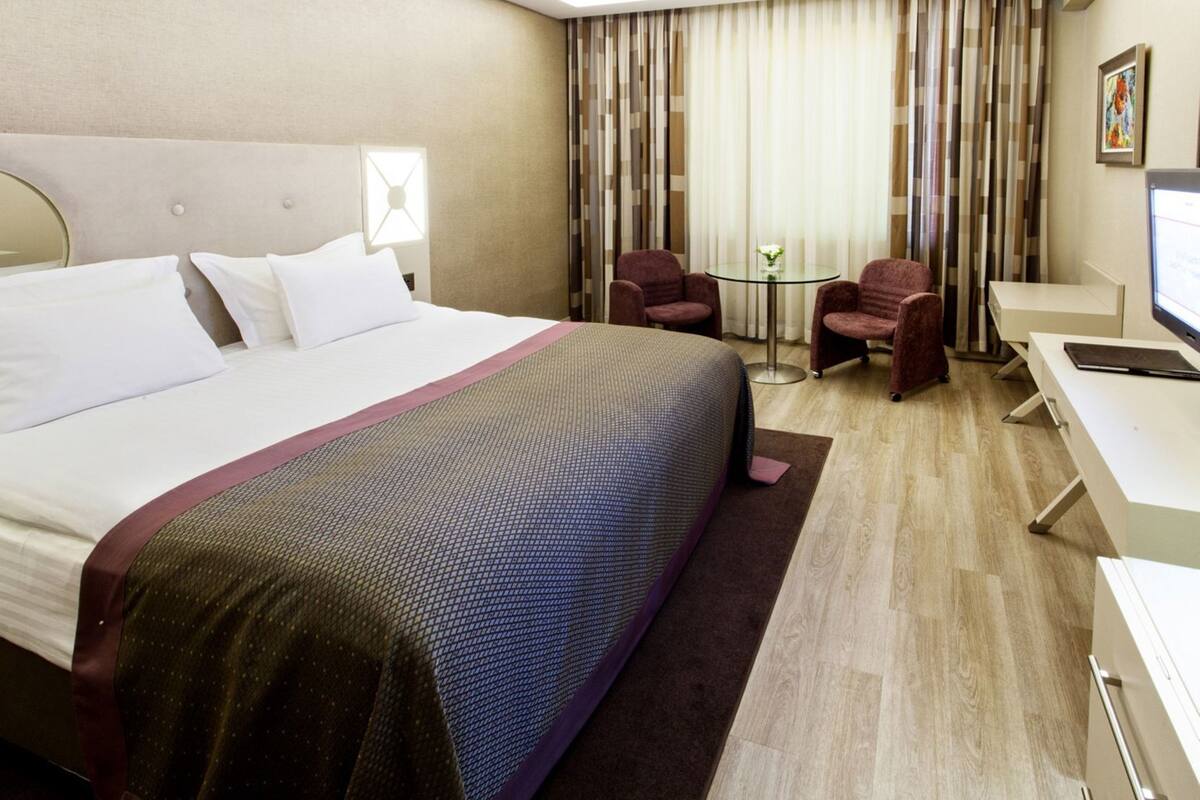 A hotel room with a large bed, two armchairs, a small table, a desk, and a television.