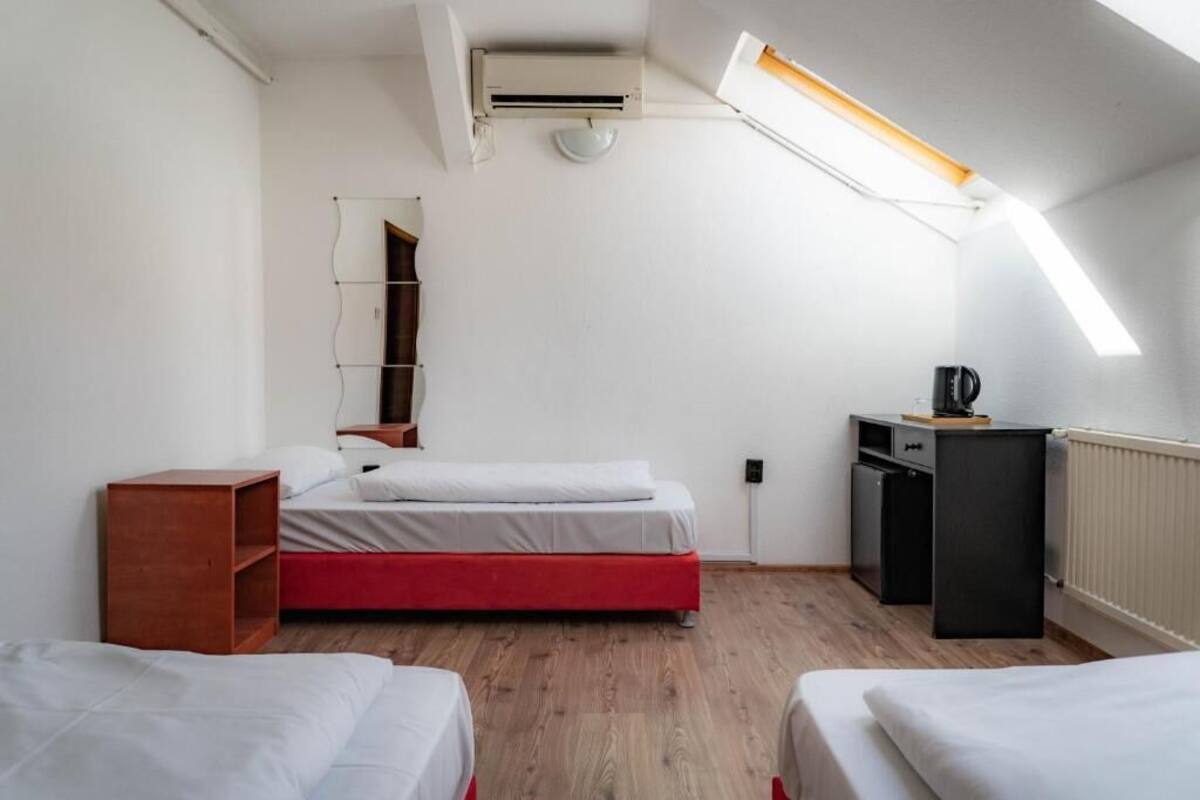 Une chambre d’hôtel de taille réduite, comprenant un lit simple, un petit bureau, un radiateur et une fenêtre avec un puits de lumière.