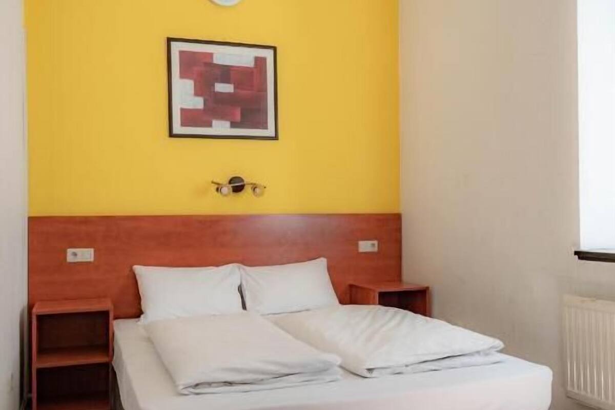 Une chambre avec un lit, une tête de lit en bois, des tables de chevet, un mur jaune et un tableau encadré.