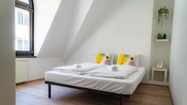 Quarto com cama, janela e uma pequena estante branca com plantas.