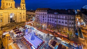 Um edifício histórico com uma cúpula imponente e edifícios adjacentes, rodeado por uma praça movimentada com bancas de mercado e uma grande árvore de Natal.