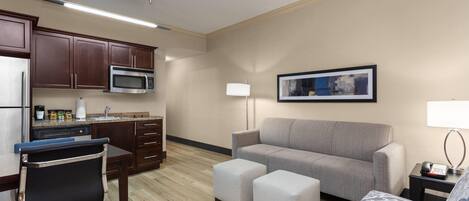 Une chambre d’hôtel moderne dotée d’une kitchenette, d’un canapé gris, de deux poufs, d’un fauteuil à motifs et d’une œuvre d’art encadrée accrochée au mur.