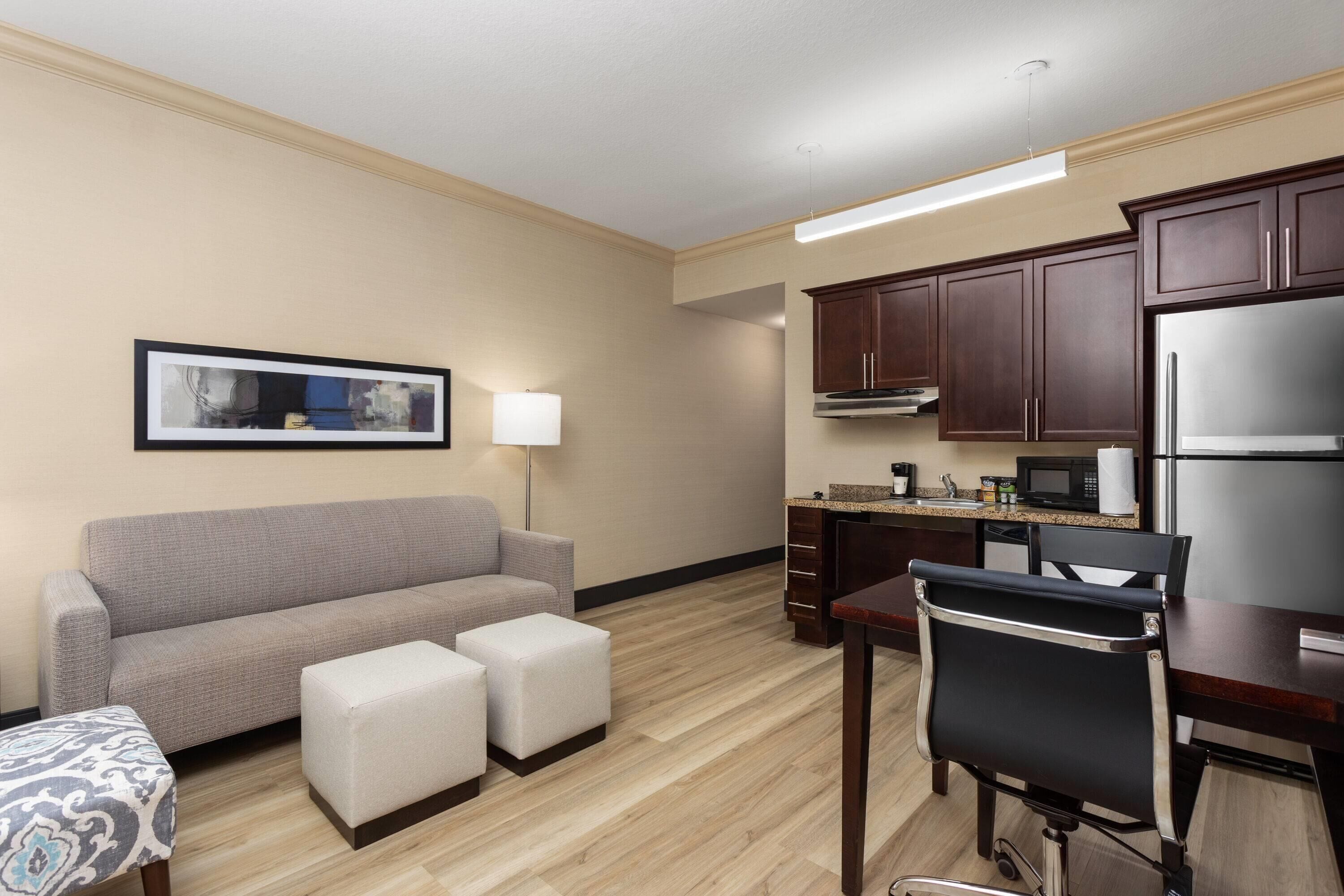 Habitación de hotel con sofá, otomanas, una mesa de comedor, una silla, zona de cocina con armarios y electrodomésticos, y un cuadro en la pared.