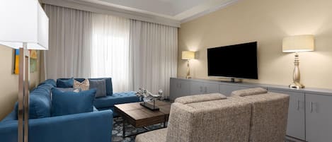 Quarto de hotel com um sofá azul, uma poltrona bege, uma televisão de tela plana e um minibar.