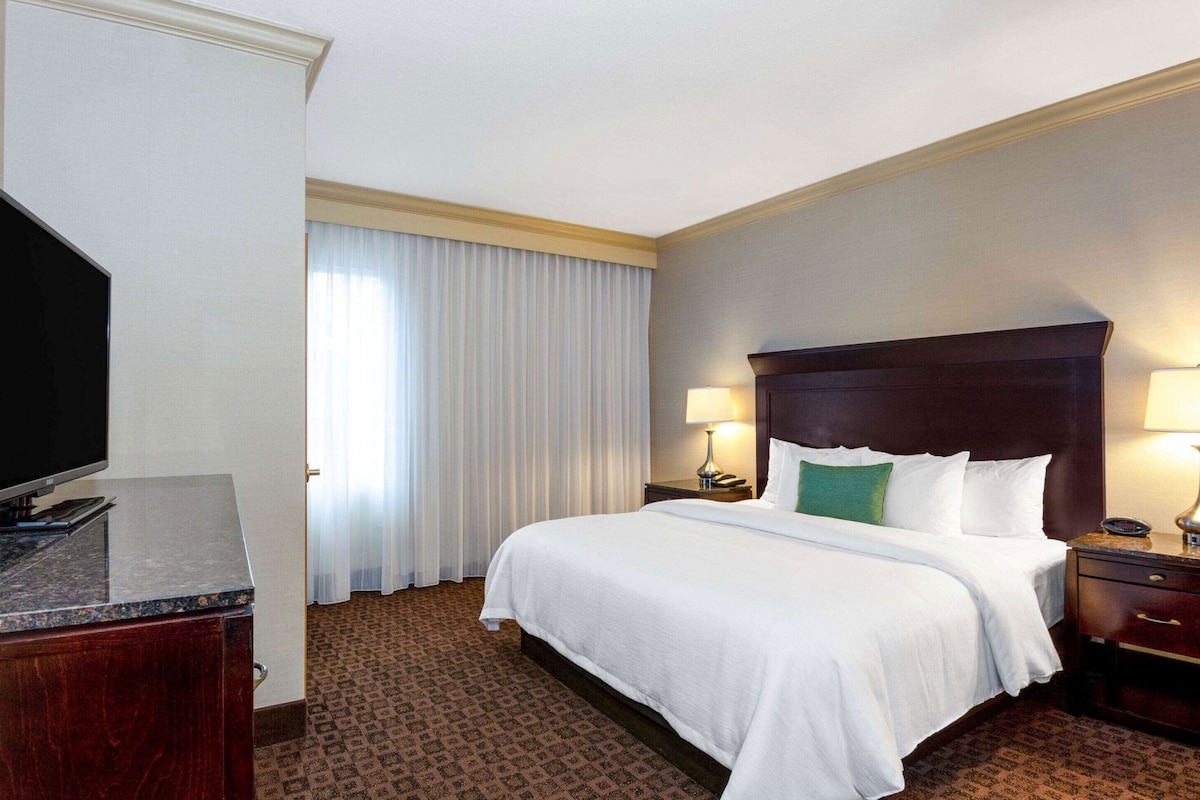 A hotel room with a bed, two bedside lamps, a television, a desk, and a window with curtains.