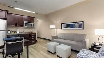 A compact living space with a kitchenette, a gray sofa, two ottomans, a patterned armchair, and a framed picture on the wall.