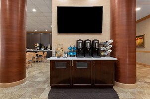 Zona de recepción de un hotel con un televisor de pantalla plana montado en la pared, una estación de café con varias máquinas de café y un área de descanso con mesas y sillas.