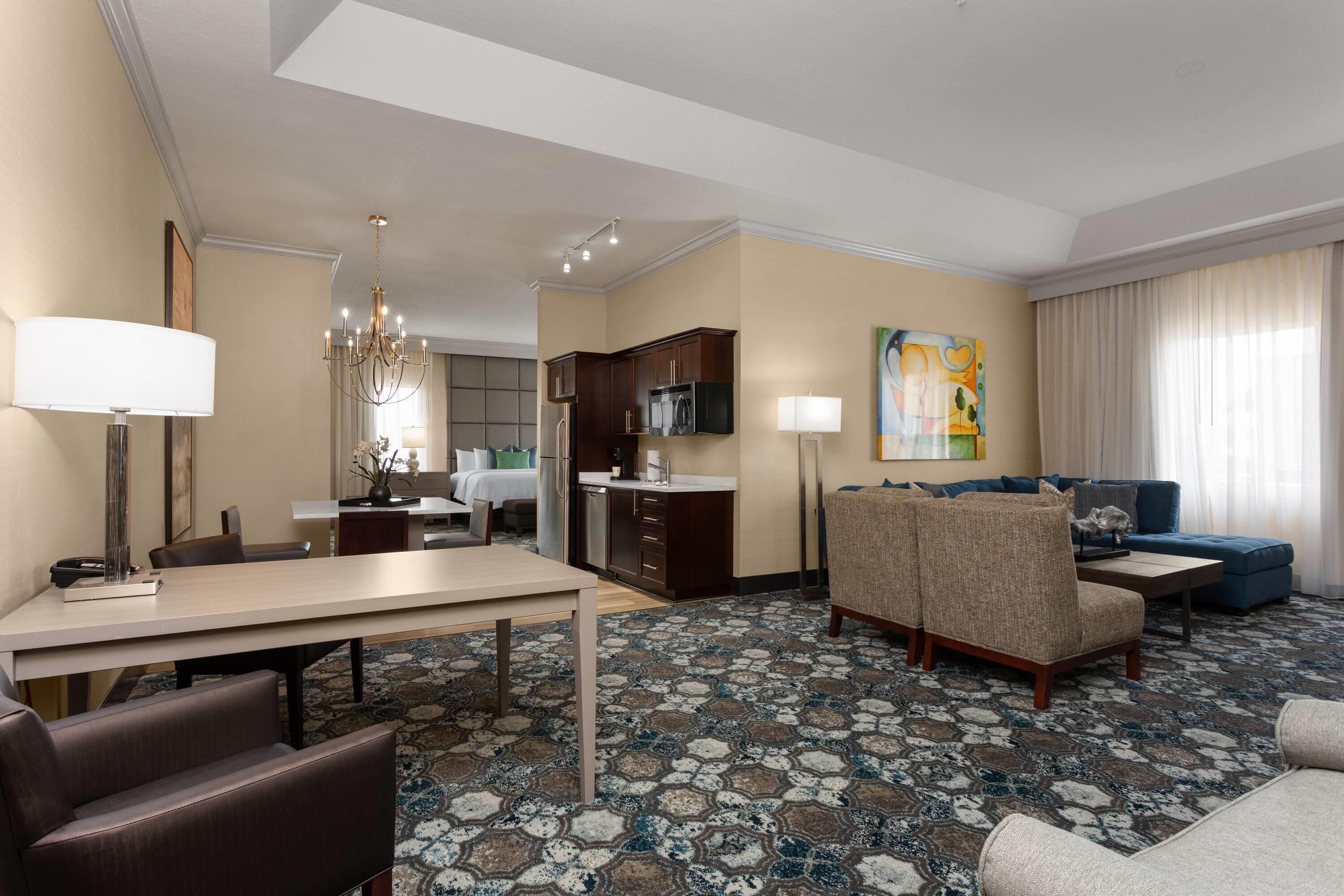 A hotel room with a dining area, a living room with a sofa and armchairs, a kitchen area, and a large window with curtains.