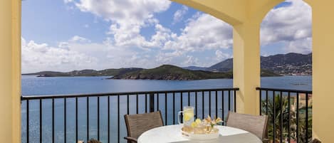 A balcony with a table and chairs, overlooking a scenic coastal view with mountains and the sea.