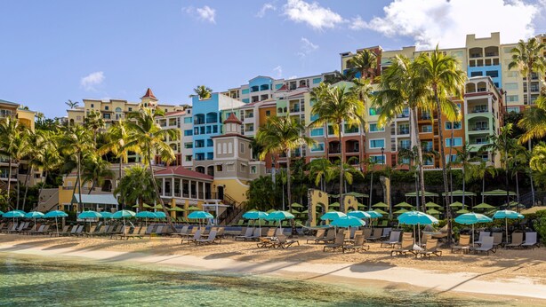 Un hotel frente a la playa con varios edificios de colores, una playa de arena y filas de tumbonas con sombrillas.