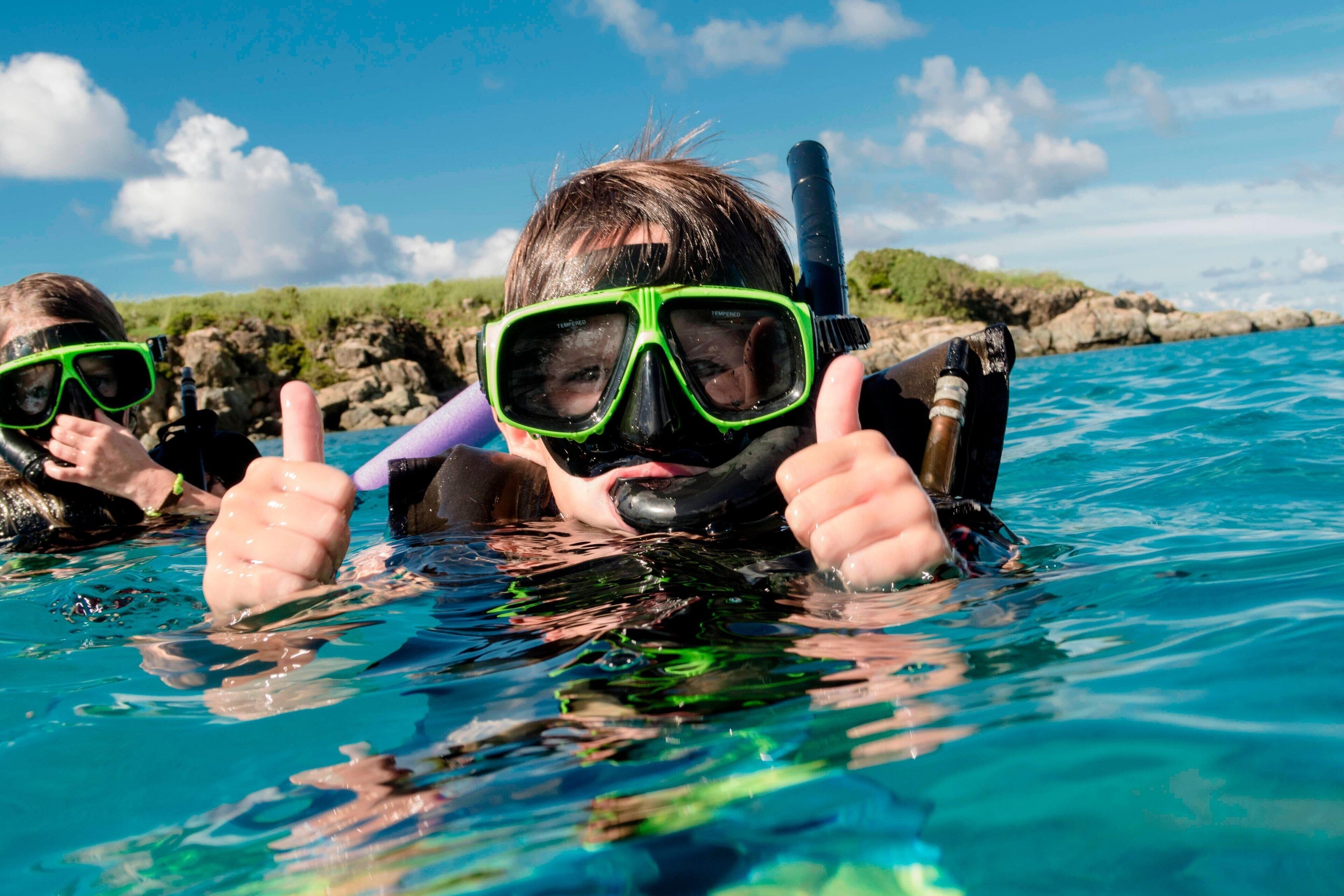Twee mensen snorkelen in helderblauw water, met rotsachtig terrein op de achtergrond.