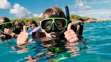 Twee mensen snorkelen in helderblauw water, met rotsachtig terrein op de achtergrond.