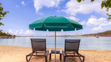 Two beach chairs under a green umbrella, a small table with a towel, and a view of the sea with boats.