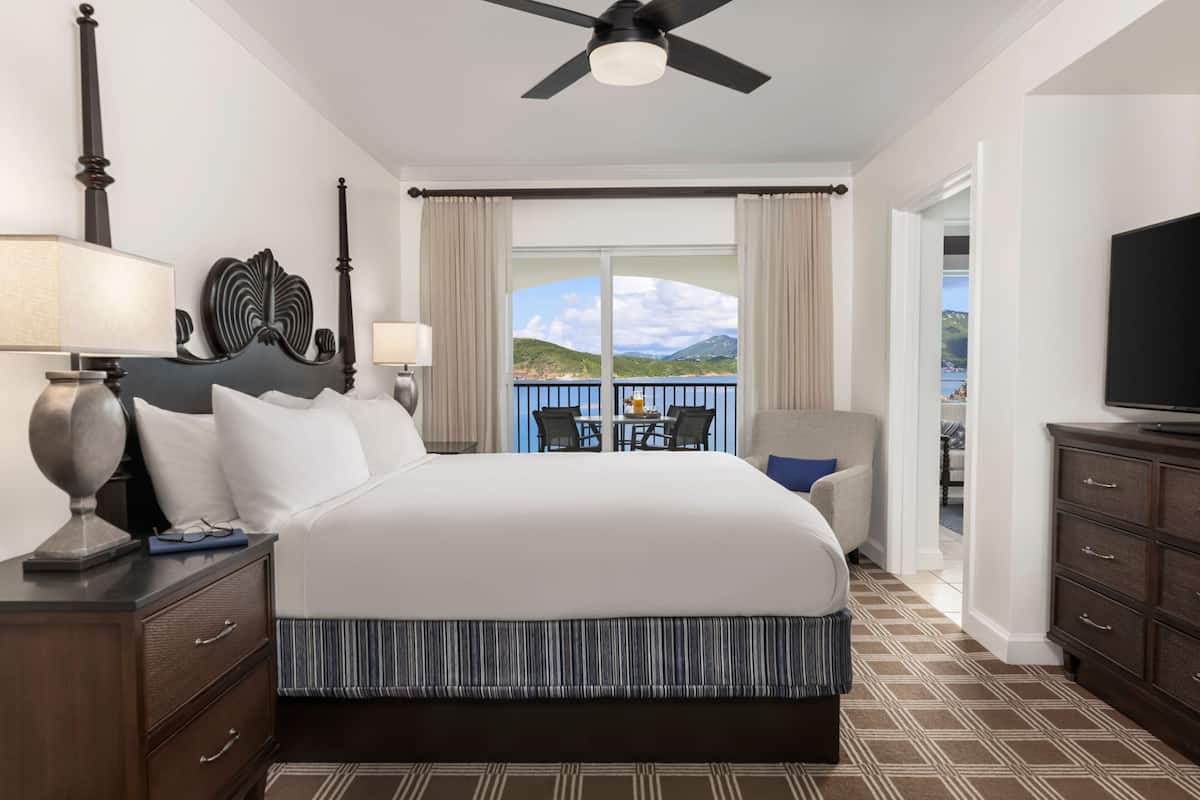 A hotel room with a large bed, a ceiling fan, a TV, a desk with a lamp, and a view of the outdoors.