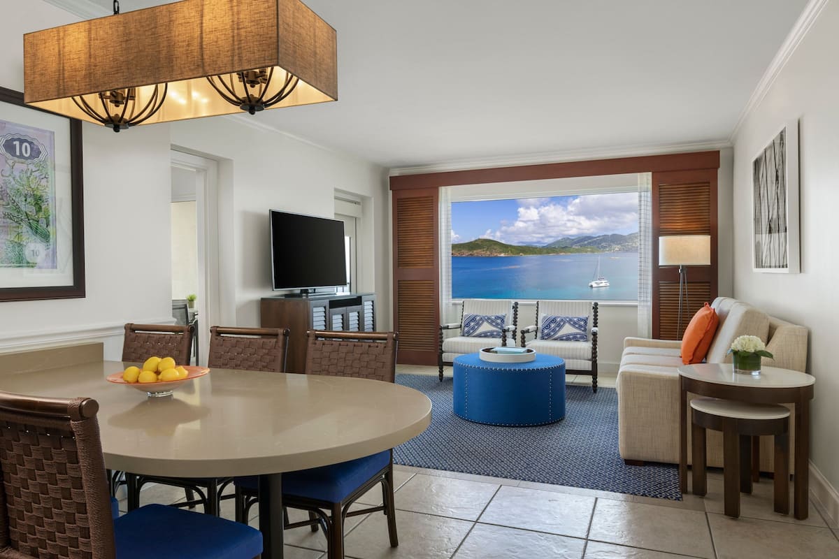 A modern living room with a dining area, a television, and a large window with a view of the sea.