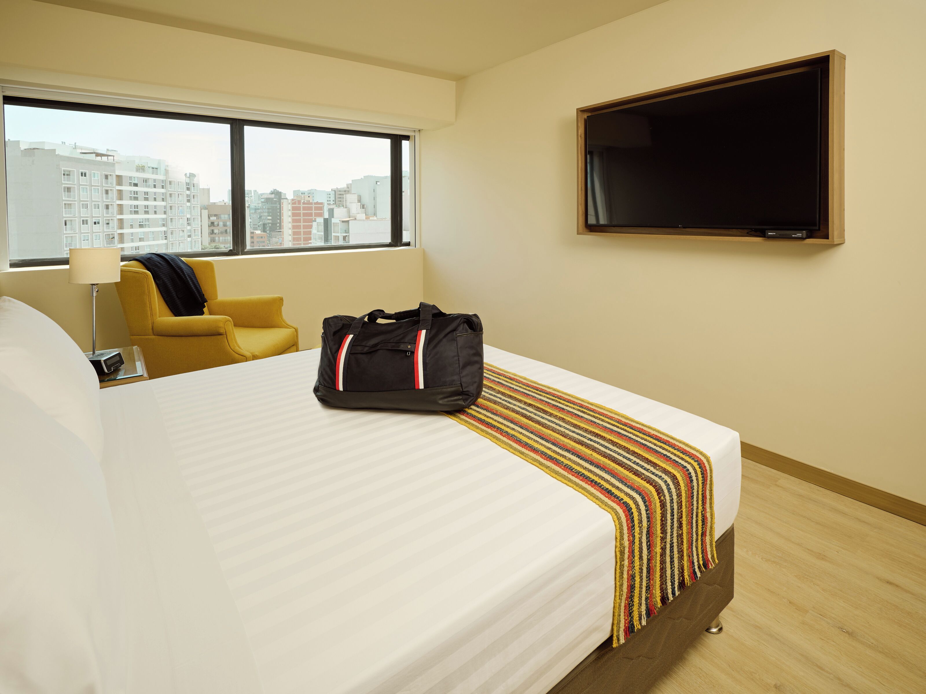 A hotel room with a bed, a TV mounted on the wall, a window with a view of buildings, a yellow armchair, and a black bag on the bed.