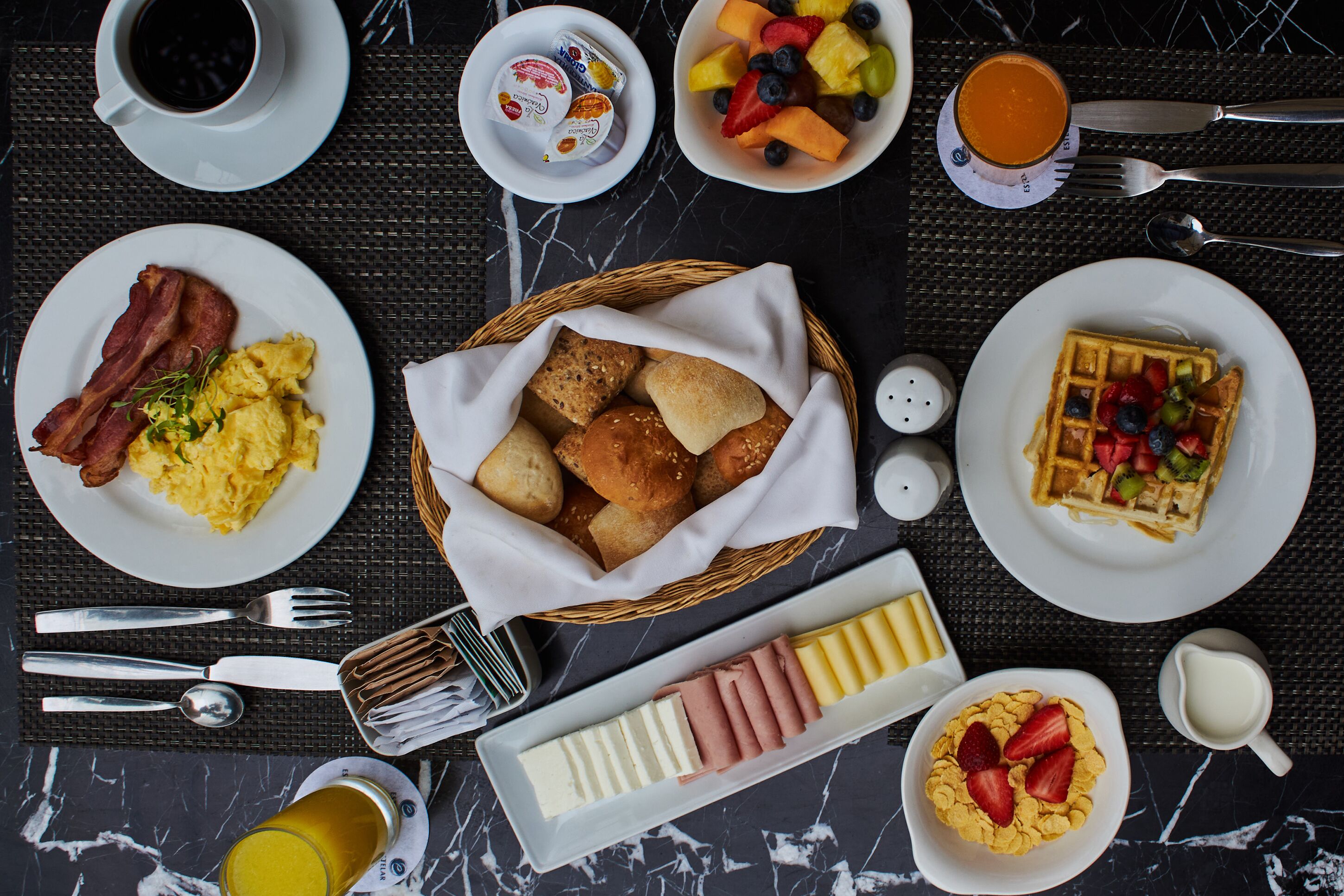 A breakfast setting with plates of eggs, bacon, waffles, and various fruits.