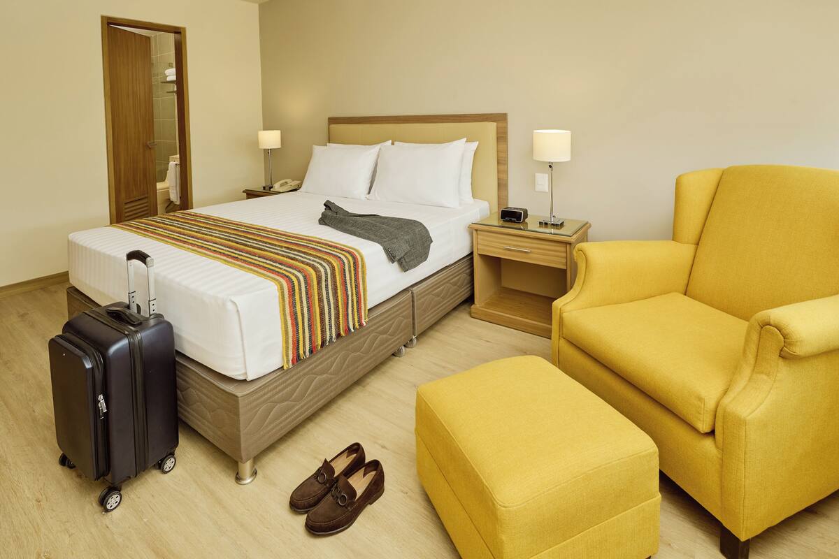 Een hotelkamer met een bed, een stoel, een nachtkastje, een lamp, een koffer en een paar schoenen.